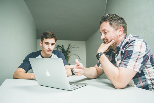 Two men smile while talking in a room with a laptop computer.