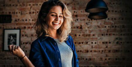 A woman in a blue denim shirt smiles in front of a brick wall.