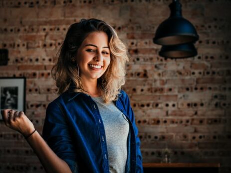 A woman in a blue denim shirt smiles in front of a brick wall.