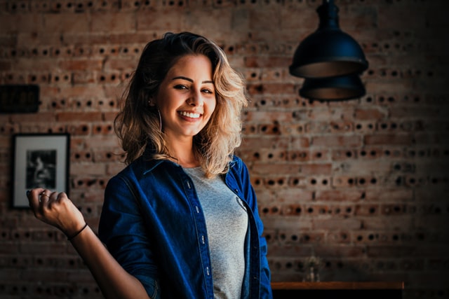 A woman in a blue denim shirt smiles in front of a brick wall.