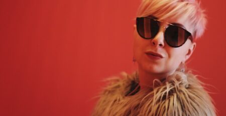 A blond woman wearing reflective sunglasses against a red background.