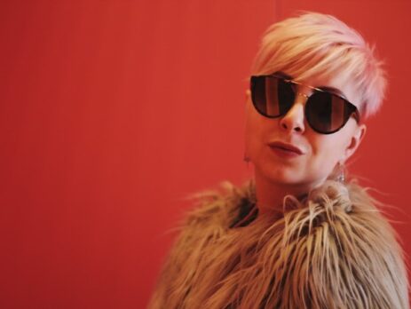 A blond woman wearing reflective sunglasses against a red background.