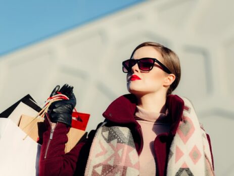 A woman in sunglasses holds shopping bags in bright sunshine.