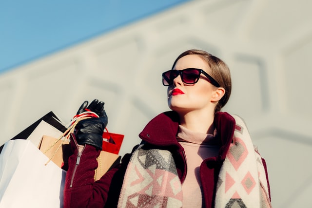 A woman in sunglasses holds shopping bags in bright sunshine.