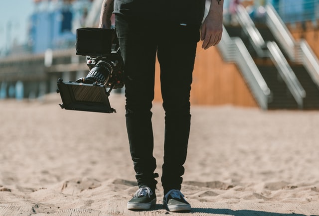 A person in Vans sneakers holds a large digital camera on sand.