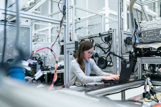 A girl works on a computer while surrounded by robots and other tech.