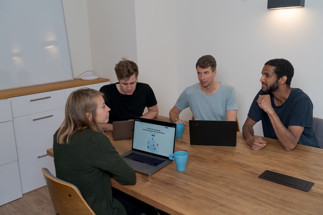 Four people sit around a brown table brainstorming with computers.