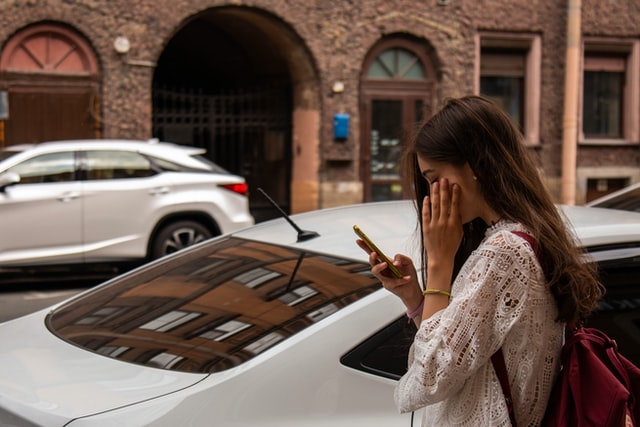 A woman in a white jacket checks her smartphone on the sidewalk near a car.