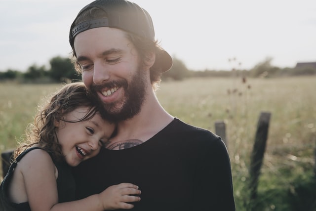 A man in a baseball cap holds a laughing child in a field.