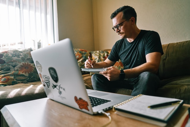 A man with brown hair and round glasses writes on a pad in front of a laptop.