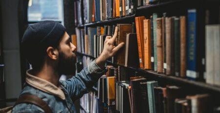 A man in a denim coat and beanie picks a book off a library shelf.