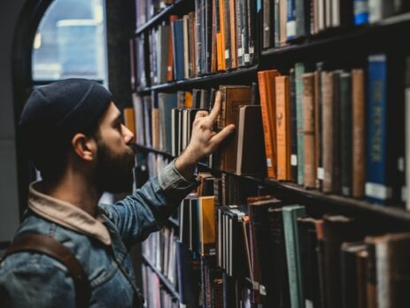 A man in a denim coat and beanie picks a book off a library shelf.
