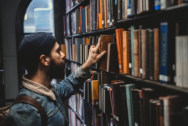 A man in a denim coat and beanie picks a book off a library shelf.