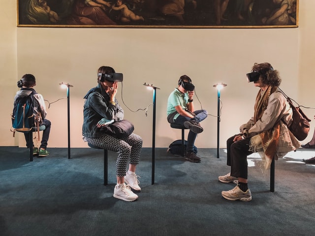 Four people sit on stools beneath a painting wearing virtual reality goggles.