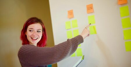 A redheaded girl in a baggy sweater smiles and points to sticky notes on a wall.