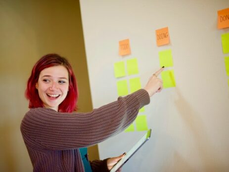 A redheaded girl in a baggy sweater smiles and points to sticky notes on a wall.