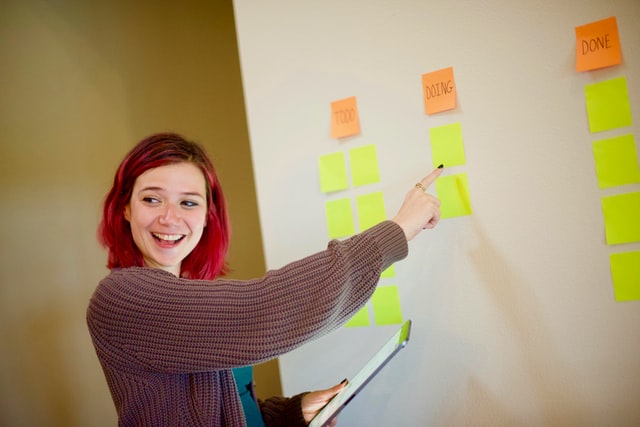 A redheaded girl in a baggy sweater smiles and points to sticky notes on a wall.