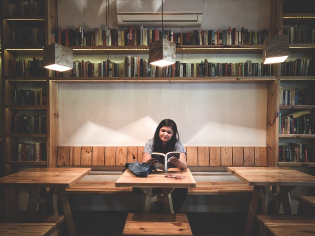 A woman with black hair reads a book in a cafe with wooden decor.