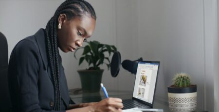 A woman in a blue sweater writes on a tablet at a desk