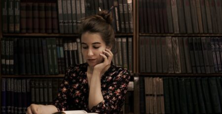 Alt-Text: A girl seated at a wooden table props her head on one hand while reading a book.