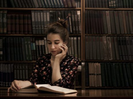 Alt-Text: A girl seated at a wooden table props her head on one hand while reading a book.