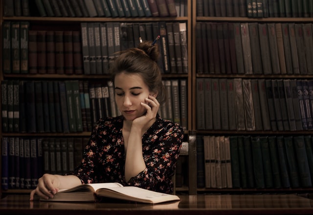 Alt-Text: A girl seated at a wooden table props her head on one hand while reading a book.