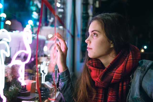 A girl peers through a glass window with neon signs.