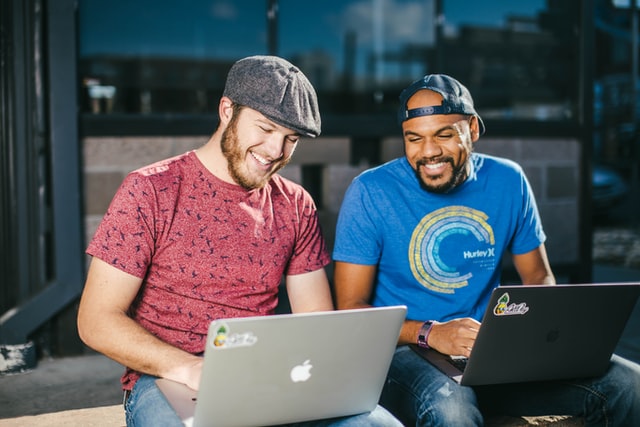 Two men sitting laughing as they work on laptop computers.