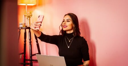 A woman with black hair takes a selfie against a wall in a pink room.