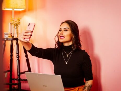 A woman with black hair takes a selfie against a wall in a pink room.