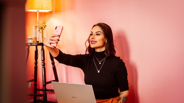 A woman with black hair takes a selfie against a wall in a pink room.