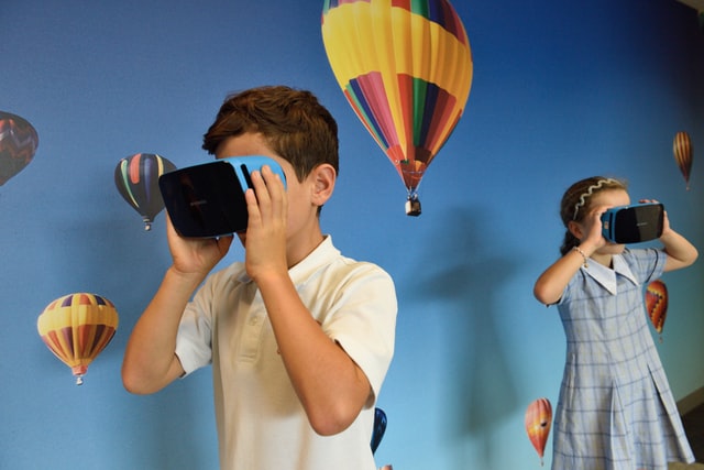 Two children use VR viewers in front of hot air balloon wallpaper.