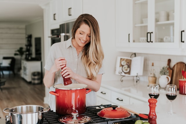 A smiling woman uses a pepper mill while cooking in a white kitchen.