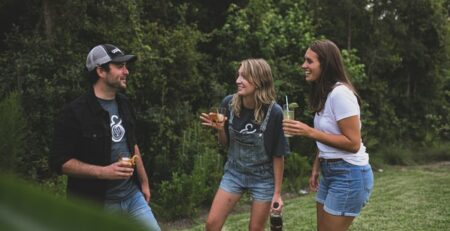 A man and two women talk while holding drinks on a lawn.