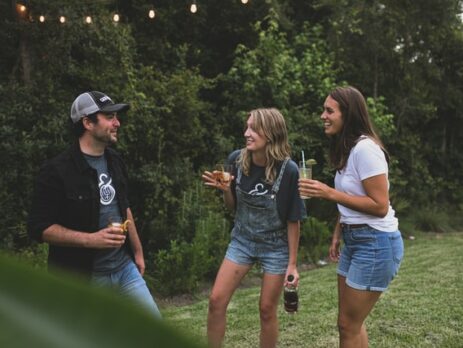 A man and two women talk while holding drinks on a lawn.