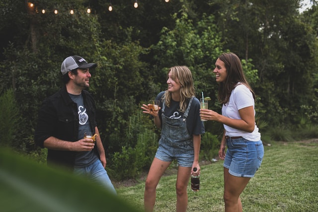 A man and two women talk while holding drinks on a lawn.