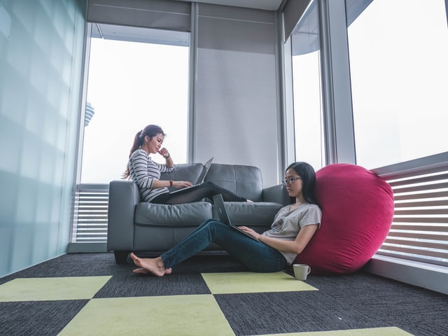 Two women work on laptops in a room with a couch.