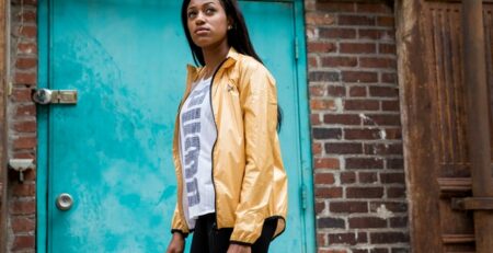 A woman in a yellow windbreaker looks skyward in front of a brick wall.