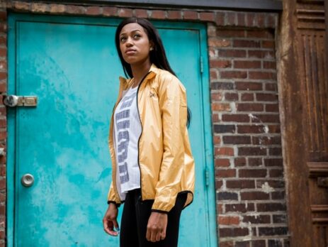 A woman in a yellow windbreaker looks skyward in front of a brick wall.