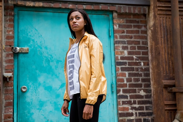 A woman in a yellow windbreaker looks skyward in front of a brick wall.