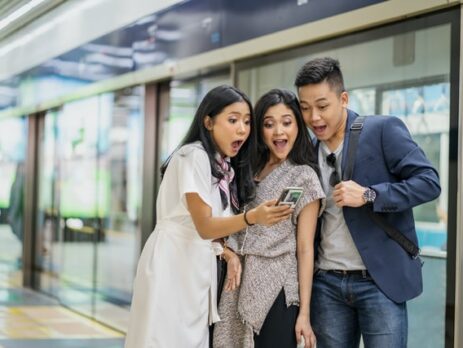 Three people act surprised as they look at a smartphone.