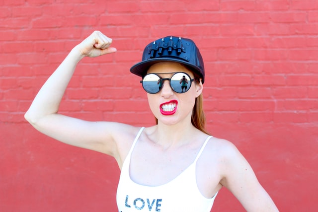 A woman in a black hat and sunglasses flexes her arm to little avail.