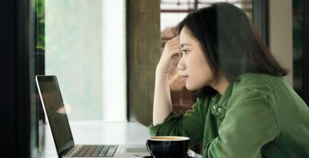 A woman wearing a green jacket looks at a laptop in a cafe.