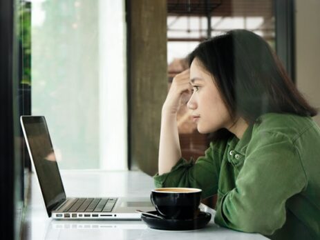 A woman wearing a green jacket looks at a laptop in a cafe.