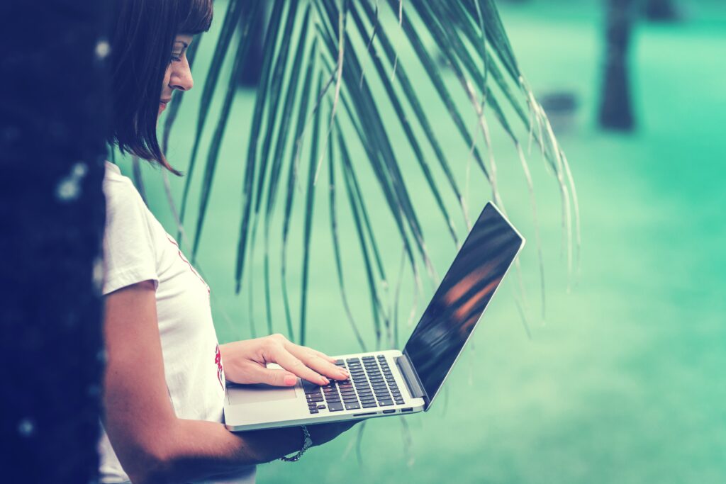 A woman in a white t-shirt uses a laptop near a tree.