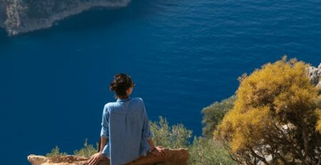A woman in a blue shirt sitting on a rock looking over a blue sea.