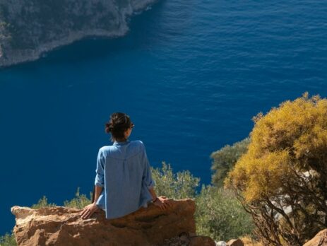 A woman in a blue shirt sitting on a rock looking over a blue sea.