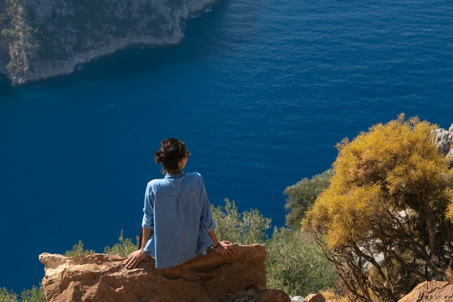 A woman in a blue shirt sitting on a rock looking over a blue sea.