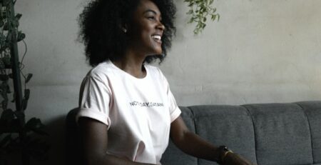 A smiling woman in a white t-shirt sitting on a grey couch.