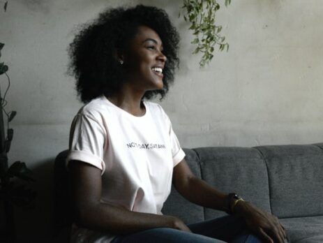 A smiling woman in a white t-shirt sitting on a grey couch.